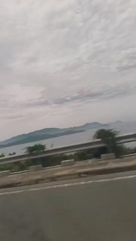 The video shows a side-view of a landscape from a moving vehicle. The sky is overcast with visible clouds. In the distance, there are several islands or hills rising from the sea. A body of water, likely the sea or a large lake, separates the islands from the foreground. A guardrail runs along the edge of the road in the midground, separating it from the natural scenery. Trees and vegetation are visible between the guardrail and the water. A lamppost stands near the road on the right side of the frame. The road itself is visible in the foreground, with white lane markings. The overall impression is a scenic drive along a coastal or lakeside route.