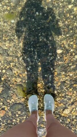 A first-person perspective video shows a person's legs and feet wading in shallow, clear water with a rocky bottom. The person is wearing white and light blue athletic shoes and red shorts. A shadow of the person is cast on the water's surface and the bottom. The person is walking forward, their feet disturbing the water and pebbles as they move.