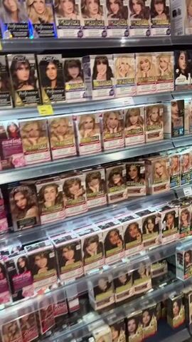 The video shows the interior of a retail store, likely a supermarket or a large drugstore, with aisles of merchandise. The camera pans across shelves stocked with various products. In the foreground, there are shelves filled with hair dye boxes, displaying images of women with different hair colors and styles. The hair dye boxes are predominantly in shades of brown, blonde, and black. As the camera moves further into the store, the shelves continue to display personal care items such as shampoos, conditioners, and other toiletries. Some shelves are also stocked with kitchenware, including frying pans and other utensils. The store appears well-lit with overhead lighting. The overall impression is of a typical retail environment offering a wide range of consumer goods. There are no people visible in the video.