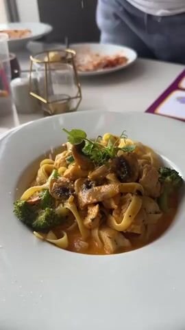 A close-up shot focuses on a bowl of pasta. A person's hand is visible in the upper right corner of the frame, sprinkling grated cheese over the dish. The pasta appears to be fettuccine noodles mixed with chicken, mushrooms, and broccoli in a creamy sauce. The dish is served on a white plate, set on a white table. In the background, another plate with pasta is visible, along with a gold-colored wire utensil holder and a person in a white shirt. The overall environment suggests a restaurant setting.