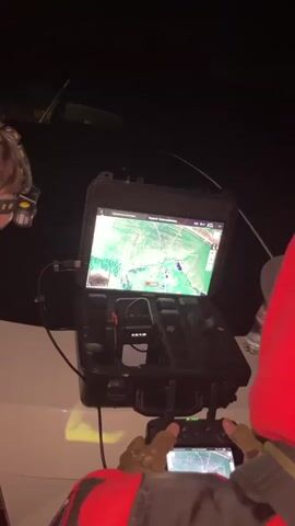 A drone is being operated at night. A person in a red jacket is holding a drone controller and looking at a tablet screen which displays a map and a live video feed from the drone. Another person wearing a headlamp is looking at a smartphone. The drone equipment is placed on the hood of a vehicle. The video feed shows a night vision image of an outdoor area with some structures and vehicles.