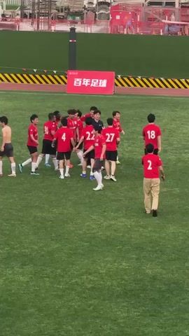 The video shows a group of people on a green sports field. Most of them are wearing red shirts with white numbers on the back. They seem to be celebrating, hugging each other, and high-fiving. One person in a similar red shirt, but with khaki pants, walks amongst them. A red banner with black and white writing is visible in the background. There is also construction and other stadium structures visible behind the banner. The camera angle is slightly elevated and captures the entire group.