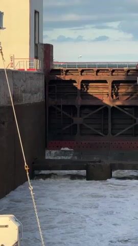 The video shows a close-up view of a lock gate on a body of water. The lock gate is made of wood and metal and appears to be old. Water is flowing through the gate, creating turbulence and white foam. The water level is rising. A concrete structure with a white building on top is to the left of the lock gate. A rope is hanging from the building and secured to something in the foreground, out of view. A bridge can be seen in the background over the lock gate. The sky is partly cloudy and the sun is setting, casting shadows on the lock gate.