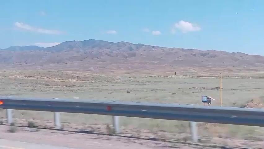 A view from a moving vehicle shows a vast, arid landscape under a clear blue sky with sparse clouds. In the foreground, a metal guardrail runs horizontally across the bottom of the frame. Beyond the guardrail, a dry, grassy plain stretches towards a distant, undulating mountain range. The mountains are a muted brown and tan, their slopes appearing rocky and barren. In the middle distance, a peculiar, weathered concrete structure stands prominently, resembling an abstract monument or ruin. The overall impression is one of a remote, desolate, and wide-open environment.
