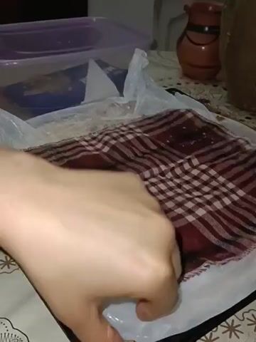 A close-up shot shows hands removing a white plastic bag, revealing a tray of baklava topped with nuts. The baklava is cut into square pieces. The tray is black, and a patterned tablecloth is visible in the background.