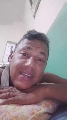 A man with dark hair and brown skin is lying on his side, propped up on his elbow, and looking directly at the camera. He is wearing a white tank top. His left arm is bent, and his hand is resting on his cheek. The man appears to be in a bedroom, as there is a pillow with a floral pattern and a blue and white striped blanket visible. In the background, a light green wall and a white air conditioning unit can be seen. There is also a framed object on the wall behind him. The lighting is dim.