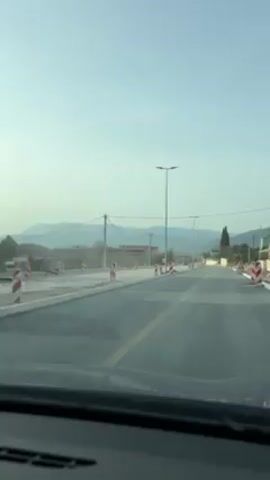A vehicle is traveling down a road, with a yellow line separating the lanes. There are red and white striped markers on either side of the road. On the left, the road has been newly paved. There are power lines stretching across the top of the frame. In the distance, mountains can be seen. Another vehicle is seen in the distance.