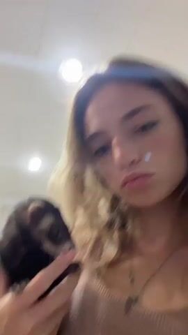 A young woman with blonde hair is holding a ferret. She is looking directly at the camera, and her expression is serious. The ferret is held close to her face, and it appears to be squirming. The background is blurry.