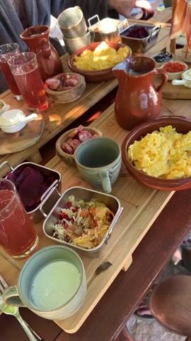 The video shows a table laden with food and drinks, likely a meal. The table is covered with wooden serving boards. There are several glasses filled with a reddish-colored drink, and at least two ceramic pitchers. Food items include what appears to be scrambled eggs or a similar dish in a clay bowl, pickled beets in a metal container, a salad with shredded vegetables and what looks like pomegranate seeds, and small dishes with various spreads and toppings like olives and red paste. There are also pieces of meat, possibly sausage or cured meat, in a small wooden bowl. A grey mug contains a pale green liquid. A person's arm, wearing a grey robe-like garment, is visible reaching for one of the glasses. The setting appears to be outdoors or in a rustic environment, with some stone-like elements in the background and natural light illuminating the scene. The camera is positioned overhead, looking down at the table.