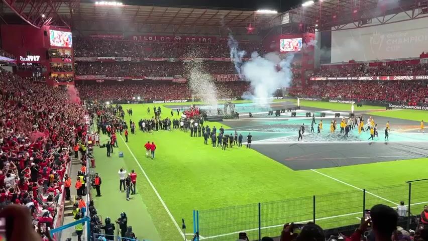 The video shows a soccer team celebrating on a platform in the middle of a field. Confetti rains down around them as fireworks shoot up from behind the platform. The field is surrounded by stands packed with cheering fans. People are filming with their phones. The field is decorated with branding for the CONCACAF Champions Cup. There are people in suits and team uniforms present on the field. Some of them are standing, some are taking pictures, and some are moving towards the center of the platform. The general atmosphere is one of celebration and excitement.