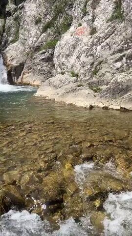 The video shows a small waterfall cascading into a clear pool of water. The pool is surrounded by rocky cliffs covered with sparse vegetation. The sky is partially cloudy.  A person briefly steps into frame on the left side of the pool before stepping out. The water is clear enough to see the rocks at the bottom of the pool.