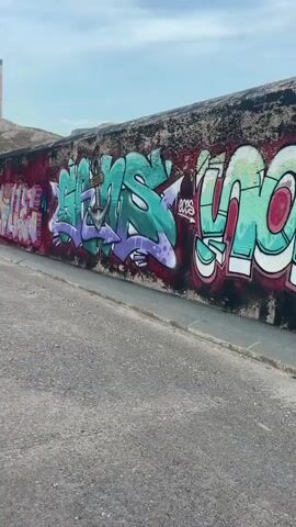 The video begins with a close-up of a graffiti-covered wall. The wall is long and appears to be made of concrete, with various colorful graffiti tags and pieces of art adorning it. The graffiti features a mix of styles and colors, including bright greens, purples, pinks, and blacks. The wall is bordered by a paved walkway and then a road. In the background, a rocky, barren hillside rises up, with a tall, thin industrial-looking structure visible at the top. As the video progresses, the camera pans to the left, revealing more of the wall and the surrounding environment. The camera then moves further away from the wall, showing the road with a white car driving away from the camera. Trees and buildings are visible on the left side of the road, and the hillside continues in the background. The sky is overcast with some light clouds. The overall scene suggests an urban or industrial area with artistic expression through graffiti.