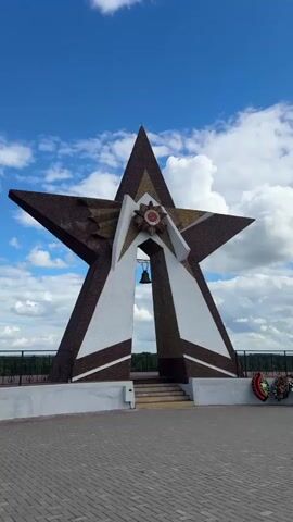 This media file features a prominent star monument under a bright blue sky with fluffy white clouds. The monument, a large, five-pointed star with a mosaic-like textured surface, stands on a paved plaza. It is a dark brown color with white accents and a golden mosaic design on the upper points. At the center of the star, there is a circular emblem with red and gold detailing and what appears to be text or symbols within it. Below this emblem, a bell hangs suspended within an opening in the monument's structure.

The monument is situated on a raised platform with a white concrete base and a decorative railing. In the foreground, the plaza is paved with light-colored rectangular bricks. To the right of the monument, a wreath with red and green elements is visible, suggesting a memorial or commemorative site. Steps lead up to the monument from a lower level. The background shows a distant treeline and more of the sky. The overall impression is one of solemnity and remembrance, likely commemorating a significant historical event. The clear blue sky and sunlight suggest it is daytime, possibly late morning or afternoon. The presence of the star and the bell strongly hints at a memorial, possibly related to military history or a significant national event. No people are visible in the frame.