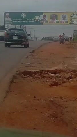 The video shows a roadside scene with a large green billboard overhead that reads "WELCOME TO THE NEW EGBA COMPUTER VILLAGE". The billboard features several portraits of men. Below the billboard, a road is visible with traffic. A black car is driving away from the camera, and another car is visible in the distance. To the right of the road, there is a dirt embankment with some loose rocks. A few people are standing near the side of the road, and one person is riding a motorcycle. The sky is overcast, and the overall impression is of a typical rural or semi-urban environment. The camera appears to be mounted on a moving vehicle, as indicated by the slight blur and the perspective.