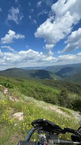 This video begins with a first-person perspective shot looking out over a vast mountainous landscape under a bright blue sky with scattered white clouds. The foreground shows the handlebars and front portion of a dirt bike, with yellow wildflowers growing on a grassy slope. The view pans slightly to the right, revealing more of the green, tree-covered mountains stretching into the distance. In the latter half of the video, the perspective shifts to show a dirt bike parked on the edge of a cliff, with a helmet resting on its handlebars. Several other dirt bikes and people are visible gathered near a large pine tree further up the slope. The overall scene depicts an outdoor recreational activity in a scenic, natural environment.