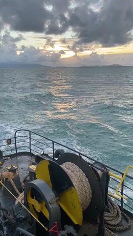 The video depicts a view from a boat deck, facing out towards a body of water under a cloudy sky with the sun peeking through. The water is slightly choppy, and some land is visible in the distance. On the deck, there is a large spool of rope and other mechanical equipment, all fixed within a metal railing.