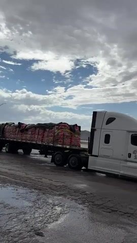 The video shows a white semi-truck with a flatbed trailer carrying several large sacks wrapped in black plastic. The truck is parked on a wet, cracked asphalt surface, possibly a roadside rest stop or shoulder. In the background, there is a highway with other trucks, green grass, and a vast, somewhat desolate landscape with rolling hills or mountains under a cloudy sky. The lighting suggests it might be overcast or late afternoon. The truck has the number 005 visible on its side. The scene appears to be in a rural or desert environment.