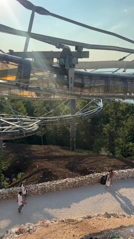 The video shows an outdoor scene with a roller coaster-like structure in the background, possibly a zip line or alpine coaster. The structure is made of metal with a transparent tube. There are people walking on a path below. The sun is shining brightly, casting long shadows. The environment appears to be a natural setting with trees and rocky terrain.