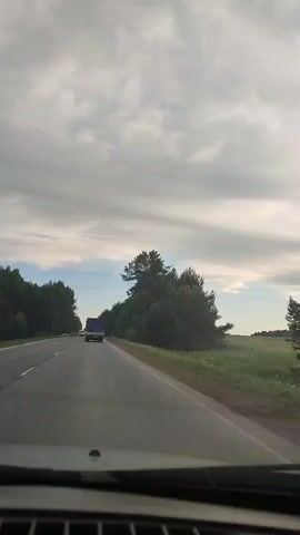 The video shows a car driving on an asphalt road. Other vehicles, including a van and a truck, are visible in the distance driving in the same direction. The road is bordered by green trees and fields on either side. The sky is overcast with grey clouds, but a bit of blue sky is visible. Music is playing in the background. The video appears to be shot from inside the car.