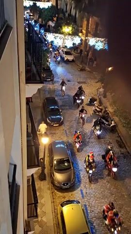 A high-angle shot shows a cobblestone street at night, illuminated by streetlights and festive overhead lighting. Several parked cars line the street, and a group of motorcyclists are riding through the scene. Most of the riders are wearing helmets, and some are dressed in holiday-themed attire. Several people are walking on the sidewalks, observing the procession of motorcycles. The camera appears to be positioned from a balcony or high window overlooking the street.