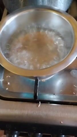 A small pot is on a gas stove with water boiling inside. The pot is silver in color and the stove is stainless steel. The burner is lit and the water is bubbling rapidly. There is some debris around the base of the pot on the stovetop. Another pot can be partially seen in the background.