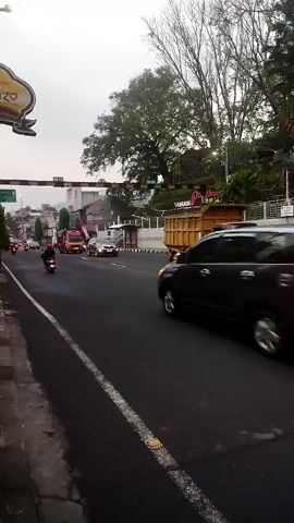 The video opens with a street scene, likely in an urban or suburban area. The time appears to be late afternoon or early evening, given the overcast sky and low light conditions. The camera is positioned at the side of a road, facing slightly uphill.

In the foreground, on the left side of the frame, is a paved sidewalk or shoulder with a white line marking the edge of the road. Further left, a portion of a yellow sign with a decorative border and some text is visible.

The road itself is paved and appears to be a multi-lane thoroughfare. Traffic is moderate, with several vehicles moving in the same direction as the camera.

On the road, a black minivan is prominently featured in the right lane, moving away from the camera. To its left, a yellow truck with a large bed is visible. Behind the truck, a white car is moving. Further in the distance, other vehicles, including motorcycles and cars, are present.

On the left side of the road, a person on a motorcycle is seen riding away from the camera.

The environment includes trees and foliage on the right side of the road, suggesting the presence of landscaping or a natural area. In the background, buildings and more trees are visible, indicating a developed area. There are also traffic signals and streetlights present.

The overall impression is a snapshot of everyday life on a busy street, with the movement of vehicles and the presence of people engaged in their daily routines.
