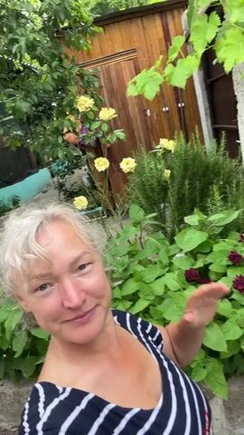 A woman with short, blonde hair is filming herself in an outdoor setting, possibly a garden or backyard. She is wearing a striped black and white shirt and is smiling at the camera. Her right arm is raised, and her hand is open as if waving. Behind her, there is a wooden structure, possibly a shed, with green foliage and yellow roses growing around it. The lighting suggests it is daytime.