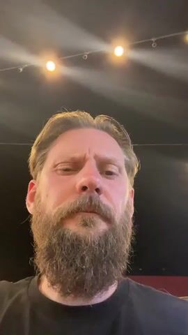 A man with a beard is looking at the camera. He has blond hair and is wearing a black shirt. The background is dark with some lights above.