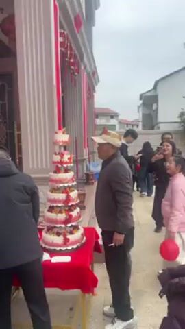 The scene unfolds outdoors, dominated by a tall, multi-tiered cake standing on a red tablecloth-covered table.  A building with red decorative elements serves as a backdrop, with people milling around. An older man wearing a hat and gray jacket walks past the cake, while a younger girl in a pink jacket looks towards it. Other individuals are scattered in the background, some looking at their phones, and one person in an apron near the building. The environment suggests a festive occasion, possibly a celebration or event. The weather appears overcast.