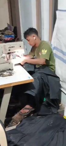A young man wearing a green t-shirt with an emblem is sitting at a sewing machine, using it to sew black fabric. He has earbuds in his ears. The sewing machine is a JUKI brand. There is a pile of black fabric on the table to the right of the sewing machine, and more black fabric is draped over his lap and the floor. He is using his hands to guide the fabric as he sews. The workspace appears to be a small, cluttered workshop with other sewing machines and materials visible in the background. The lighting is artificial. He is wearing sandals.