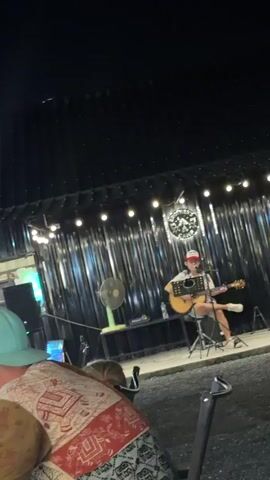 The video shows a person playing an acoustic guitar and singing into a microphone on a stage at night. The stage has a black curtain backdrop with string lights. A logo for "BROTHER'S CAMP" is visible above the performer. To the left of the stage, there is a fan and some audio equipment. The foreground shows the backs of audience members.
