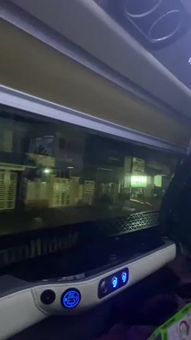 The video shows the interior of a vehicle, likely a bus, looking out a window at night. The window reflects interior lights and some of the vehicle's dashboard, which has illuminated blue buttons and a digital display. Outside the window, there are buildings with illuminated signs and what appear to be stacked pallets. The scene outside is dark and wet, suggesting it may be raining. The vehicle appears to be in motion, as evidenced by the slight blurring of the outside scenery. There are no people clearly visible in the video.