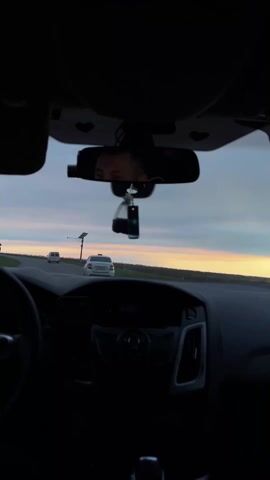 The image provides a first-person perspective from the interior of a car, looking out through the windshield onto a road. The car's dark dashboard occupies the lower half of the frame, showing the steering wheel partially on the left and the central console with various controls. A smartphone is mounted on the dashboard to the left.

In the center, the rearview mirror prominently reflects the driver's eyes and forehead, showing a neutral and focused expression. A dark, liquid-filled air freshener or car accessory hangs from the rearview mirror. Above, the car's interior ceiling is dark, with subtle heart-shaped cutouts visible on the sun visor clips.

Outside, the asphalt road stretches ahead, appearing straight or with a gentle curve. Two other vehicles are visible in the distance: a white sedan, possibly a Ford Focus, followed by a white van further ahead. To the right of the road, a modern utility pole with a solar panel array and a light fixture stands. The landscape beyond the road consists of low green fields or natural vegetation.

The sky exhibits the pastel hues of either dawn or dusk, with soft orange and pink light near the horizon gradually transitioning into light blue and grey overhead, indicative of the "golden hour." The weather appears clear with good visibility. The scene suggests a calm drive on an open, intercity, or rural highway, consistent with the provided location of Novodonetskaya, Russia. No visible text is legible in the image.
