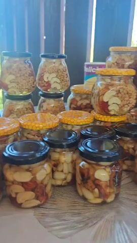 The video shows a collection of glass jars filled with nuts and dried fruits. Some jars have black lids, while others have lids with a honeycomb pattern. The jars are arranged on a surface, possibly a table, with a wooden fence or wall in the background. The lighting suggests an outdoor or semi-outdoor setting. There are no people visible in the video.