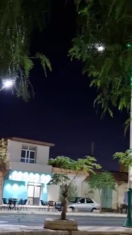 The video captures a nighttime scene on a street. A street lamp is illuminated, casting a glow. Trees with dark green foliage hang into the frame from the top. In the background, a light-colored building with a blue awning is visible, suggesting it might be a shop or cafe. Several tables and chairs are set up outside the establishment. To the left, a large orange garbage truck is parked. A white car is parked on the right side of the street. The sky is dark, indicating it is nighttime. The overall ambiance is that of a quiet street scene after dark.