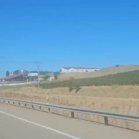 The video shows a rural landscape under a clear blue sky. In the foreground, a highway guardrail runs along the bottom of the frame. Beyond the guardrail, dry, grassy terrain is visible. In the middle ground, there are several buildings, including a large building with a reddish-brown roof and a distinctive dome-shaped structure with a metallic appearance. Another larger white building with a reddish roof is situated on a hill in the background. There are also power lines and poles present. The scene appears to be a sunny day, possibly in a warm climate. There are no people visible in the video.