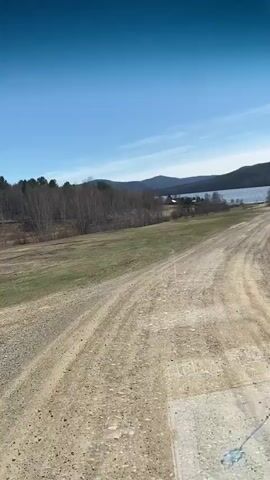 The video shows a dirt road leading toward a lake, framed by green fields on either side. The road is uneven and appears rocky, with tire tracks visible. In the distance, a lake stretches across the landscape, surrounded by tree-covered hills. The sky is clear blue with a few wispy clouds. Bare trees are visible on the left side of the road. The perspective suggests the video was taken from a moving vehicle.