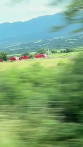 The video, set in Pevtsite, Bulgaria, captures a red train, branded "ÖBB", partially derailed in a lush green, hilly landscape during the daytime. Several red carriages or locomotives are visible off the tracks, resting in a grassy field. The footage is filmed from a moving vehicle, resulting in significant motion blur in the foreground. In the background, rolling mountains can be seen under a cloudy sky.