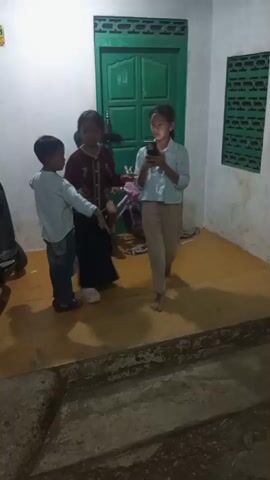 A group of children are gathered in a dimly lit room. In the foreground, a child in a light blue shirt and beige pants is standing, holding a phone. To their left, a child in a maroon jacket and dark skirt is also standing, seemingly interacting with the child holding the phone. A younger child in a light blue shirt and blue jeans is standing to the left of the other two, looking towards the child with the phone. In the background, a green door is visible, as well as a window with a patterned screen. A motorcycle is parked to the left of the group. The floor appears to be a light yellow tile, and the walls are white. Another child, partially obscured, is visible in the background on the right. The children appear to be engaged in some sort of activity or game.