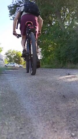 A low-angle, rear-facing video shows a person riding a bicycle on a gravel path. The person is wearing a backpack and dark shorts. Trees and foliage line the right side of the path, while a white fence and several parked cars are visible on the left. The sky is clear and blue. The camera remains stationary, capturing the cyclist's movement away from the viewer.