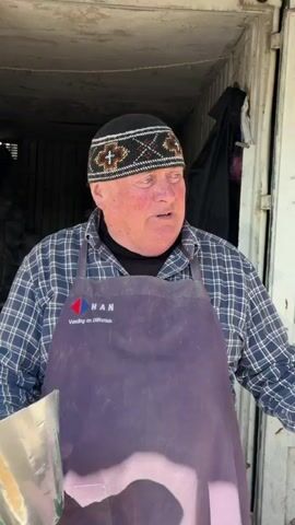 A man in a cap and apron stands in front of several sacks of grain. He is wearing an apron that says "HAN Voeding en diëtetiek," as well as gloves. He is standing in a doorway. A scoop is in one of the sacks. The sacks contain corn, wheat, and oats.
