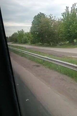 The video shows a view from a moving vehicle looking out the window at a road. The road is gray and has grass and trees along the side. There is a metal guardrail running parallel to the road. The sky is overcast. As the vehicle moves, it approaches a fork in the road and passes a small building surrounded by trees. The building has a green sign on it.