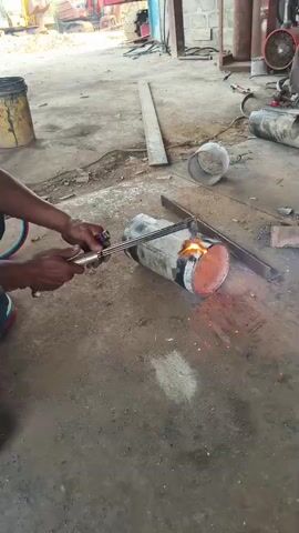 A person is shown cutting a metal pipe with a torch. The pipe is lying on the ground in an industrial setting. Sparks are flying from the point of contact between the torch and the pipe. The person is wearing gloves and protective clothing. In the background, there are various tools and equipment, including a yellow bucket, a red construction vehicle, and a large gas cylinder. The ground is dirt and concrete.