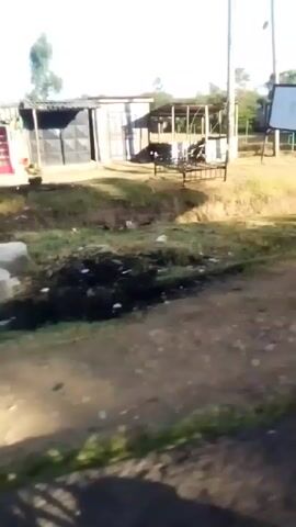 The video shows a view from a moving vehicle traveling through a rural area. The road is dirt and unpaved, and the environment is mostly dry and dusty. There are several simple structures visible, including small shops and houses made from corrugated iron and wood. A large water tank and a blue building are also visible. The sky is clear and blue, indicating it is daytime. There is minimal activity, although some individuals are visible near the structures.