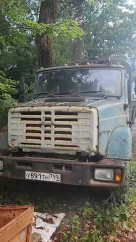 A medium shot focuses on the front of an old, light blue truck parked on the side of a dirt road. The truck has a weathered appearance, with rust and peeling paint visible on its grille and body. A license plate with Russian characters is attached to the front bumper. The truck is surrounded by lush green foliage and trees, indicating an outdoor setting. In the background, a person rides a bicycle on the dirt road, passing by the truck. The video appears to be a steady, unedited recording of the truck and its surroundings.