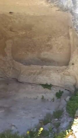 The video shows a person's perspective exploring what appears to be an abandoned quarry or a historical site. The initial frames are dark and show close-ups of rough, rocky cave-like structures with some vegetation and debris on the ground. The camera then pans to reveal a more open outdoor environment with a path leading through a rocky area. There are remnants of stone structures, a wooden walkway with a railing, and various types of vegetation, including grass and trees. The sky is visible with clouds, suggesting an outdoor setting during the daytime. There are no people visible in the video.
