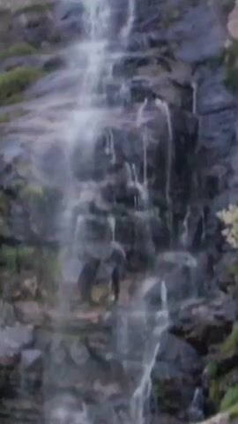 A close-up shot of a waterfall cascading down a rocky cliff. The rocks are dark and wet, with patches of green moss and grass growing in crevices. The water is white and foamy, creating a misty spray as it falls. The bottom of the waterfall is not visible.