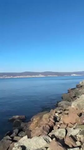 The scene captures a tranquil seascape under a clear, bright blue sky. The water, a deep azure, stretches towards a distant landmass, which is marked by a faint outline of hills and structures along the coast. The foreground is dominated by a rocky shoreline composed of medium-sized, irregularly shaped stones that blend shades of gray, brown, and dark red. The rocks appear stable and grounded, creating a rugged edge between land and sea.