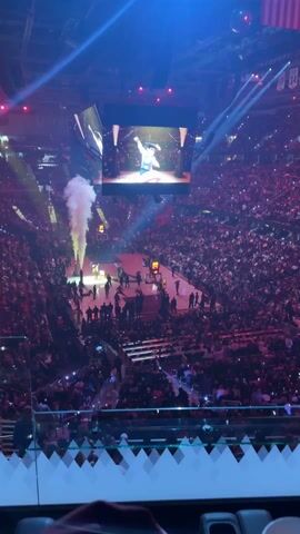 Here is a description of the video:

The video shows a large basketball arena, presumably before a game. The arena is packed with people. A large video screen hangs above the center court, showing images of people and cityscapes. There are bursts of flames and plumes of smoke erupting from various points on the court. Spotlights scan the arena, adding to the spectacle. People are milling about on the court. The video is shot from an elevated position, looking down into the arena.