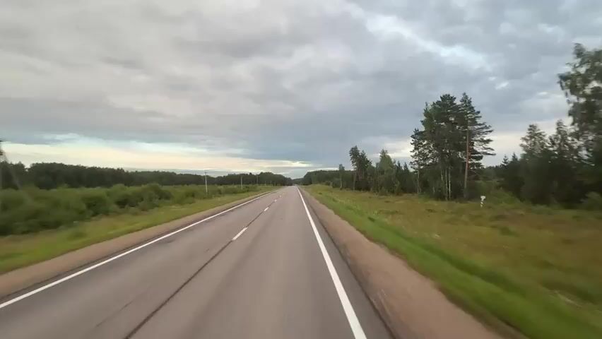 A first-person view from a vehicle traveling on a two-lane asphalt road. The road has white painted lines marking the lanes and a white line along the right edge. The vehicle is traveling through a rural landscape with dense green trees and vegetation on both sides of the road. The sky is overcast with gray clouds. The time appears to be late afternoon or early evening based on the lighting conditions.